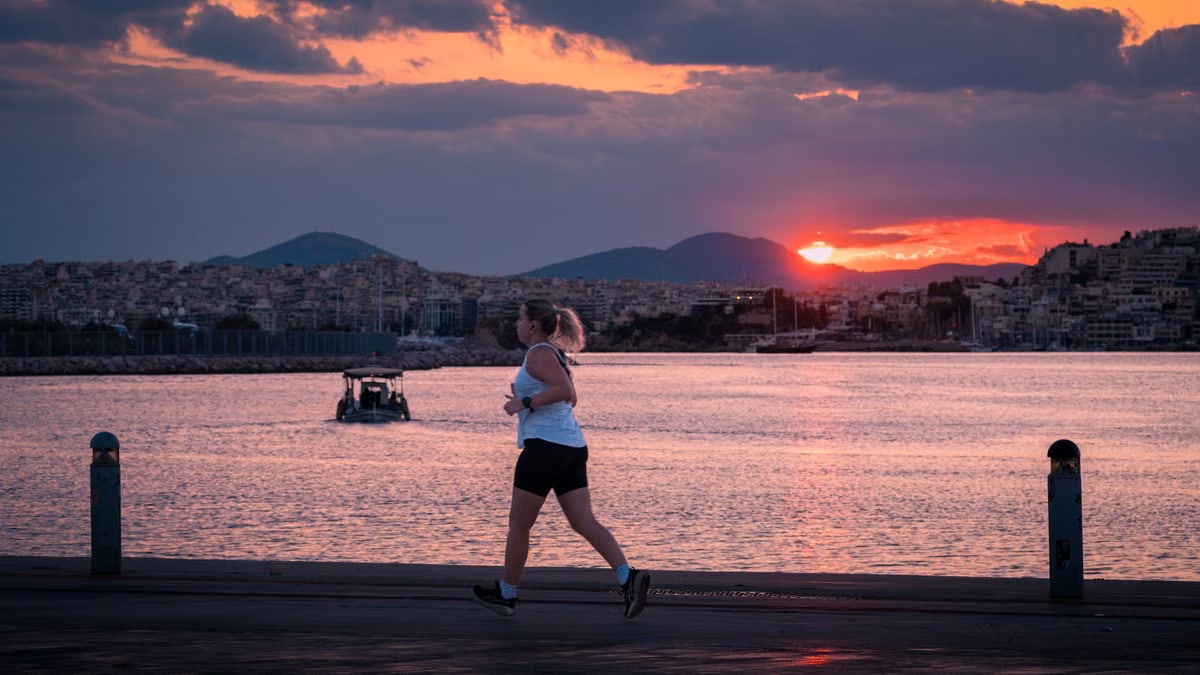 A woman jogs along the waterfront at sunset with a cityscape and boat in the background.